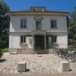 Ein zweistöckiges weißes Gebäude mit einem zentralen Eingang und mehreren Fahrrädern, die auf dem Kopfsteinpflaster davor abgestellt sind, umgeben von Bäumen und Sträuchern.