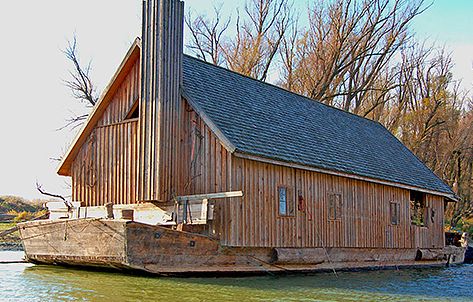 Ein großes hölzernes Wassermühlenhaus mit schrägem Dach liegt an einem Fluss, umgeben von blattlosen Bäumen im Hintergrund.
