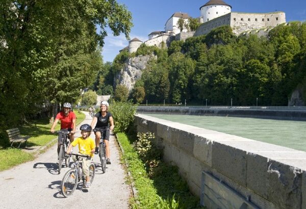 Drei Personen und ein Kind fahren mit dem Fahrrad auf einem gepflasterten Weg neben einem Fluss, mit einem bewaldeten Hügel und einem großen Schloss im Hintergrund.