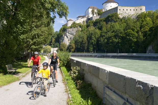 Drei Personen und ein Kind fahren mit dem Fahrrad auf einem gepflasterten Weg neben einem Fluss, mit einem bewaldeten Hügel und einem großen Schloss im Hintergrund.