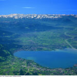 Luftaufnahme eines großen Sees, umgeben von grünen Feldern und Städten, mit entfernten Bergen unter einem klaren blauen Himmel.