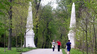 Drei Personen gehen auf einem gepflasterten Weg durch einen grünen Park, vorbei an zwei hohen weißen Steinobelisken, die von Bäumen umgeben sind.