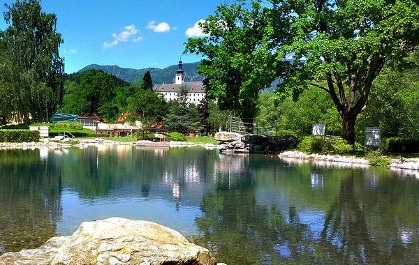 In einem ruhigen, von Felsen und Bäumen umgebenen Teich spiegelt sich ein entferntes weißes Gebäude mit einem Turm unter einem klaren blauen Himmel.