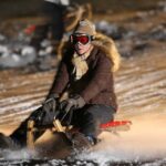 Eine Person in Winterkleidung und mit Schutzbrille fährt nachts auf einem Holzschlitten einen verschneiten Hang hinunter, wobei der Schnee um sie herum aufspritzt.