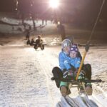 Zwei Personen fahren nachts mit einem Schlitten einen verschneiten Hang hinauf, wobei sie sich an einem Liftgriff festhalten, während im Hintergrund andere Schlittenfahrer und Lichter zu sehen sind.