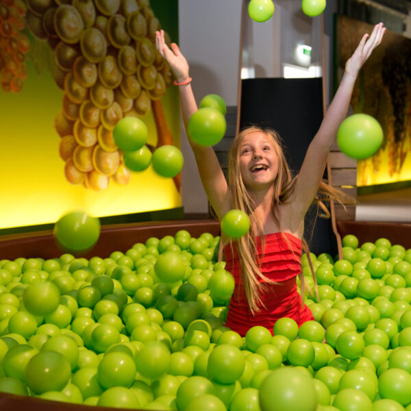 Ein Mädchen in einem roten Kleid wirft grüne Plastikbälle, während es in einer großen, mit grünen Bällen gefüllten Grube sitzt.
