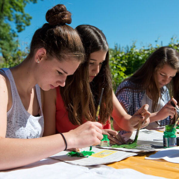 Drei junge Frauen sitzen an einem Tisch im Freien und beschäftigen sich unter sonnigem Himmel mit grünen Farben und Pinseln mit Malprojekten.