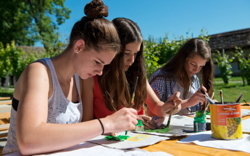 Drei junge Frauen sitzen an einem Tisch im Freien und beschäftigen sich unter sonnigem Himmel mit grünen Farben und Pinseln mit Malprojekten.