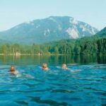 Drei Personen schwimmen in einem See, der von Wäldern und Bergen umgeben ist, unter einem klaren Himmel.