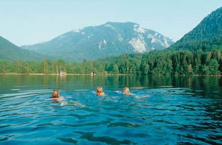 Drei Personen schwimmen in einem See, der von Wäldern und Bergen umgeben ist, unter einem klaren Himmel.