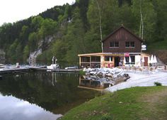 Ein hölzernes Gebäude am See mit Sitzgelegenheiten im Freien steht am ruhigen Wasser, umgeben von Bäumen und einem Hügel im Hintergrund.