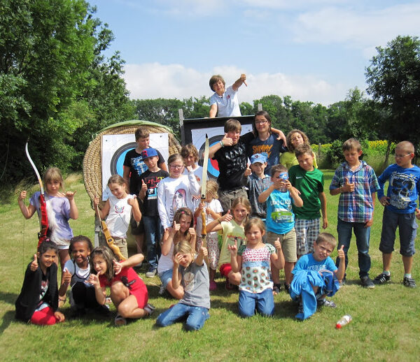 Eine Gruppe von Kindern posiert an einem sonnigen Tag im Freien vor Zielscheiben für das Bogenschießen, einige halten Spielzeugschwerter und zeigen die Daumen nach oben.