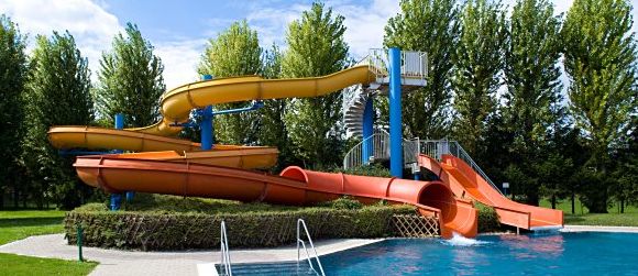 Zwei bunte, orangefarbene und gelbe Wasserrutschen führen von einer Metallplattform hinunter in ein blaues, von Bäumen umgebenes Außenschwimmbecken.