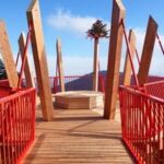 Eine hölzerne Aussichtsplattform mit rotem Geländer und hohen Holzbalken, die eine bewaldete Landschaft unter blauem Himmel überblickt.