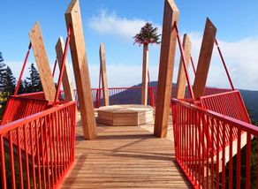 Eine hölzerne Aussichtsplattform mit rotem Geländer und hohen Holzbalken, die eine bewaldete Landschaft unter blauem Himmel überblickt.