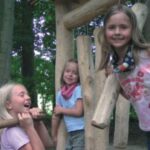 Drei junge Mädchen spielen und lächeln auf einem hölzernen Klettergerüst in einem Park mit Bäumen im Hintergrund.