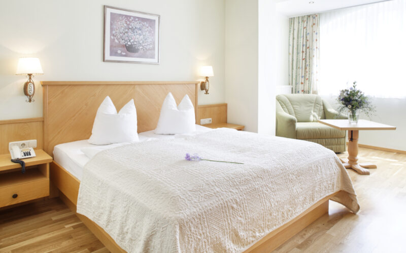 Ein ordentlich gemachtes Hotelbett mit weißer Bettwäsche und Kissen, ein kleiner Tisch mit Stühlen am Fenster und eine Blume auf der Bettdecke in einem hellen, aufgeräumten Zimmer.