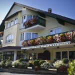 Ein dreistöckiges Haus mit Balkonen und Fenstern, die mit bunten Blumenkästen und grünen Geländern geschmückt sind, unter einem klaren blauen Himmel.