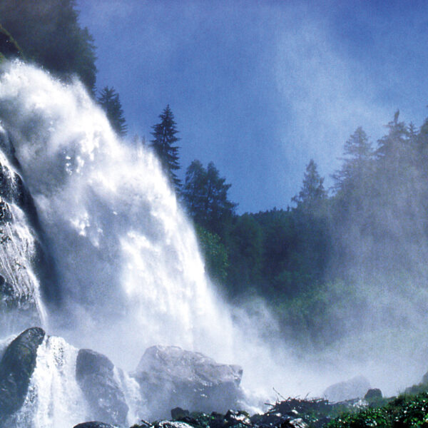 Ein Wasserfall stürzt über felsige Klippen, die von immergrünen Bäumen umgeben sind, unter blauem Himmel in die Tiefe, wobei Nebel aus dem fallenden Wasser aufsteigt.