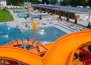 Blick auf einen Freibadkomplex mit leuchtend orangefarbenen Wasserrutschen, Becken, Liegeflächen und entspannten Menschen.