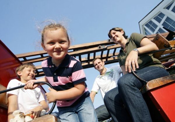 Drei Kinder und zwei Erwachsene sitzen lächelnd auf einer Holzbank im Freien unter strahlend blauem Himmel, im Hintergrund ist ein Gebäude zu sehen.