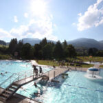 Freibad mit Menschen, die schwimmen und sich entspannen, umgeben von Grasflächen, Bäumen und Bergen unter einem sonnigen Himmel.