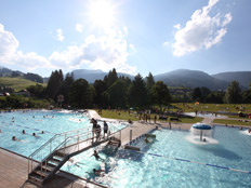 Freibad mit Menschen, die schwimmen und sich entspannen, umgeben von Grasflächen, Bäumen und Bergen unter einem sonnigen Himmel.