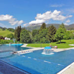 Außenschwimmbad mit Springbrunnen, umgeben von grünen Rasenflächen, Bäumen und Hügeln unter einem teilweise bewölkten Himmel.
