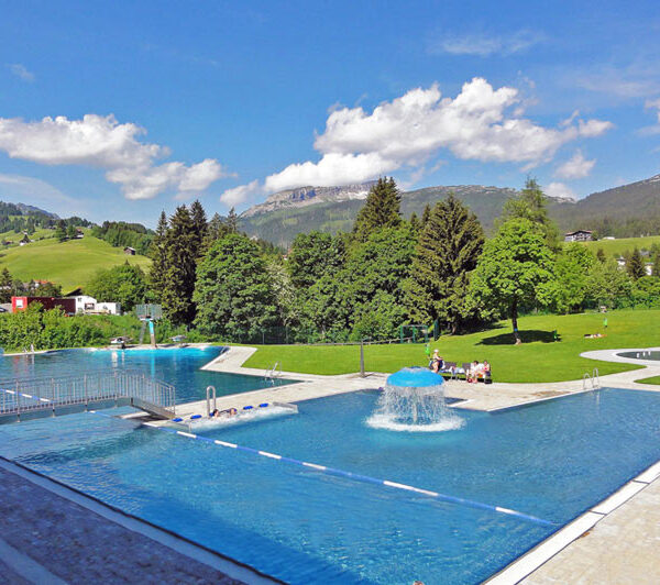 Außenschwimmbad mit Springbrunnen, umgeben von grünen Rasenflächen, Bäumen und Hügeln unter einem teilweise bewölkten Himmel.