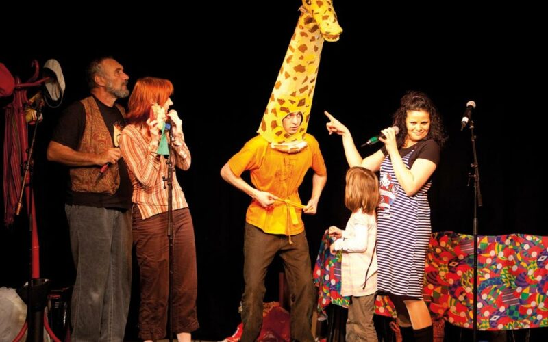 Fünf Personen treten auf der Bühne auf; eine trägt ein Giraffenkostüm, andere stehen oder halten Mikrofone, und im Hintergrund steht ein bunter Tisch.