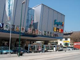 Modernes Gebäude mit Metallverkleidung, großen Glasfenstern und mehreren Plakaten an der Außenseite, unter denen Autos geparkt sind.