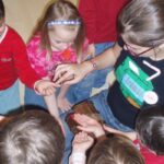 Eine Gruppe kleiner Kinder versammelt sich um sie und streckt ihre Hände aus, um etwas zu berühren oder zu betrachten, das ein Mädchen mit Brille und schwarzem T-Shirt in der Hand hält.