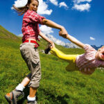 Zwei Kinder spielen draußen auf einer Wiese, wobei das eine das andere an den Armen herumwirft, unter einem blauen Himmel mit vereinzelten Wolken.