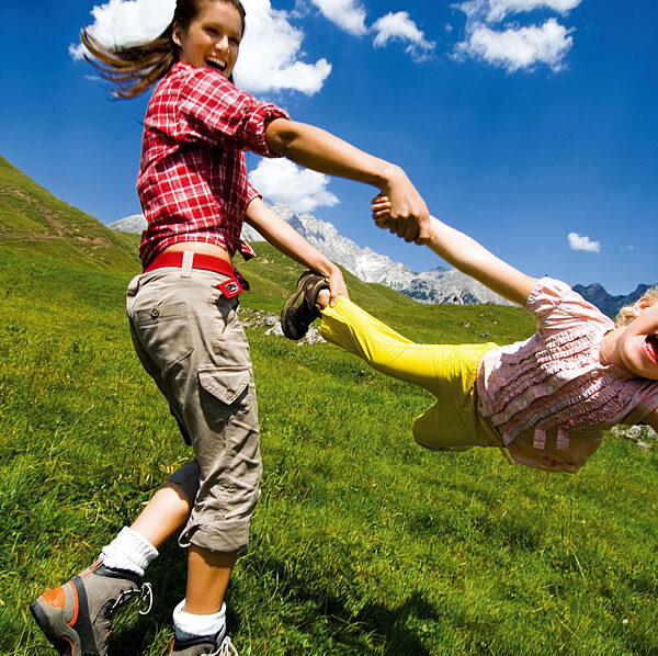 Zwei Kinder spielen draußen auf einer Wiese, wobei das eine das andere an den Armen herumwirft, unter einem blauen Himmel mit vereinzelten Wolken.