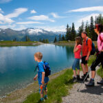 Eine vierköpfige Familie in Wanderkleidung steht an einem klaren Bergsee mit schneebedeckten Gipfeln und Bäumen im Hintergrund an einem sonnigen Tag.