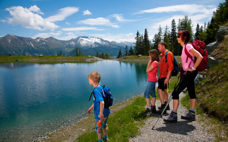 Eine vierköpfige Familie in Wanderkleidung steht an einem klaren Bergsee mit schneebedeckten Gipfeln und Bäumen im Hintergrund an einem sonnigen Tag.