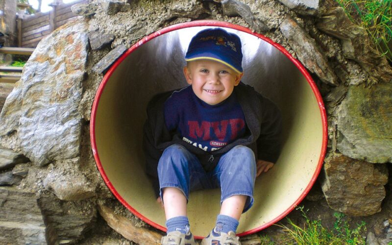 Ein kleiner Junge mit blauer Mütze und Turnschuhen sitzt in einem runden Spielplatztunnel aus Stein und Metall und lächelt in die Kamera.
