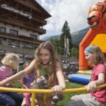 Vier Kinder spielen auf einem Karussell vor einem Gebäude, mit einer orangefarbenen, tigerförmigen Hüpfburg im Hintergrund auf einer Wiese.