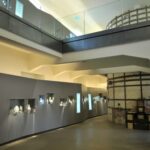 Ein modernes Museumsinterieur mit Glasvitrinen an der Wand, in denen verschiedene Artefakte ausgestellt sind, und einer freiliegenden, runden Backsteinstruktur im Hintergrund.