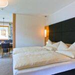 Helles Hotelzimmer mit einem Doppelbett, gepolstertem Kopfteil, Tisch und Stühlen am Fenster und moderner Beleuchtung.