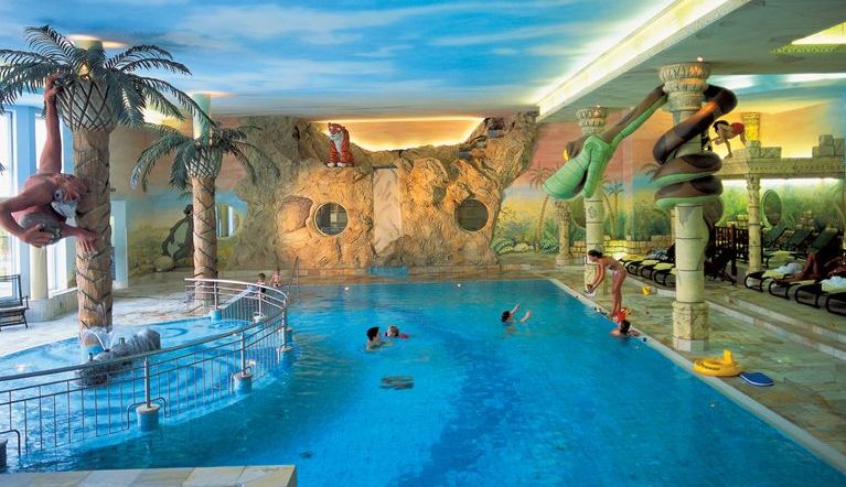 Hallenbad mit tropischem Dschungel-Thema, mit Rutschen, bemalten Wänden, Palmen-Dekorationen und Kindern, die im Wasser spielen.