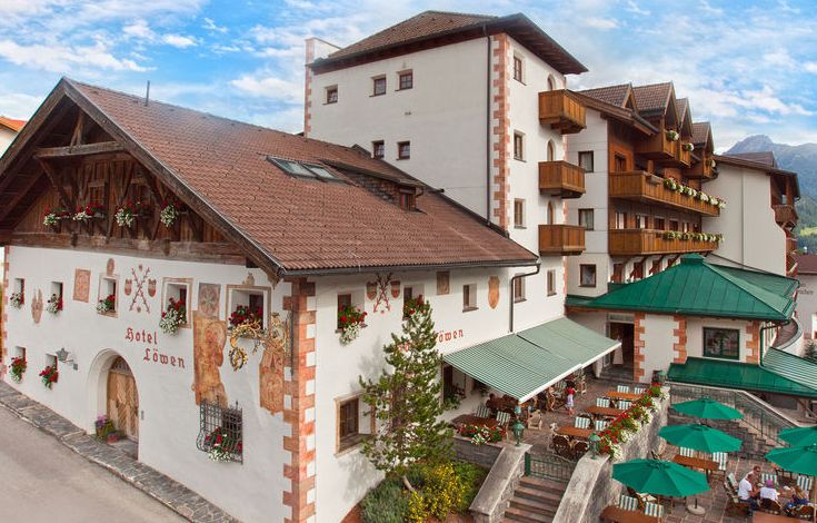 Ein traditionelles Alpenhotel mit weißen Wänden, Holzbalkonen, Blumenkästen und einer Terrasse mit grünen Sonnenschirmen vor einem teilweise bewölkten Himmel.