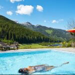 Eine Person schwimmt in einem Außenpool mit Bergen und Wäldern im Hintergrund, während ein paar Leute unter Sonnenschirmen auf dem Pooldeck sitzen.