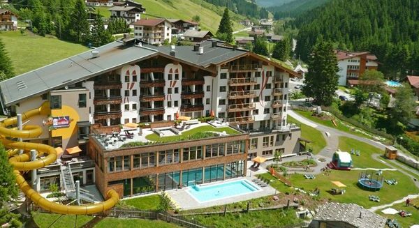Ein großes Gebirgsresort mit Wasserrutsche, Freibad, Spielplatz und grünen Hügeln inmitten eines Alpendorfs.