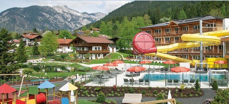 Alpine Ferienanlage mit Gebäuden im Chalet-Stil, Außenpool, roten Sonnenschirmen, gelber Wasserrutsche, Spielplatz und Berglandschaft im Hintergrund.