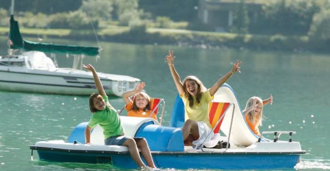 Vier Personen, zwei Erwachsene und zwei Kinder, sitzen auf einem Tretboot auf einem See, lächeln und winken mit erhobenen Armen. Im Hintergrund sind ein Segelboot und Grünflächen zu sehen.