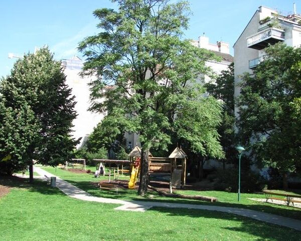 Ein kleiner Spielplatz mit einer Rutsche und einem Klettergerüst befindet sich inmitten von Bäumen und Gras, mit Wohnhäusern im Hintergrund.