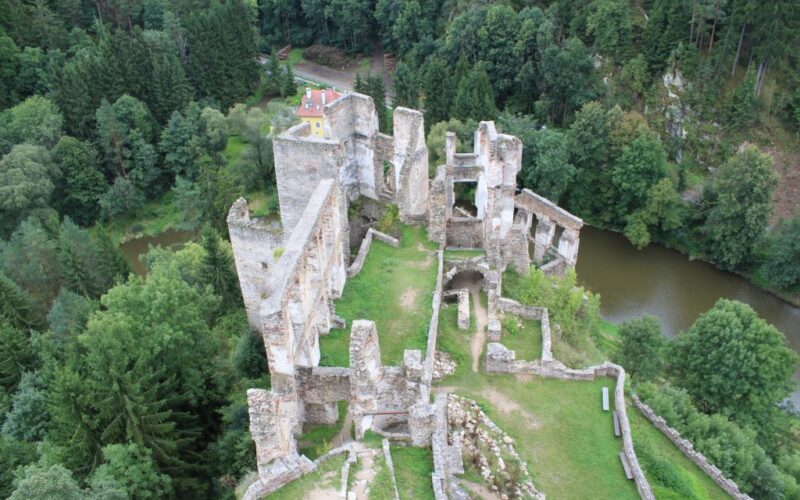 Luftaufnahme einer steinernen Burgruine inmitten eines dichten grünen Waldes, mit einem Fluss in der Nähe und einem kleinen Haus im Hintergrund.