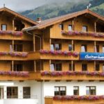Ein Alpenhotel aus Holz mit mehreren Balkonen, die mit Blumenkästen geschmückt sind, vor einer Kulisse aus grünen Hügeln und Bäumen. Auf den Schildern steht "Galtenberg.