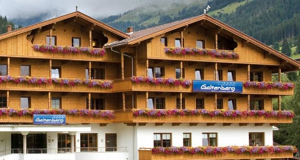 Ein Alpenhotel aus Holz mit mehreren Balkonen, die mit Blumenkästen geschmückt sind, vor einer Kulisse aus grünen Hügeln und Bäumen. Auf den Schildern steht "Galtenberg.